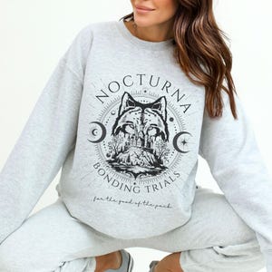 May include: Light gray sweatshirt with a black wolf graphic and the text "NOCTURNA BONDING TRIALS" with a crescent moon design. The phrase "for the good of the pack" is printed below the graphic. The sweatshirt is part of a matching set.