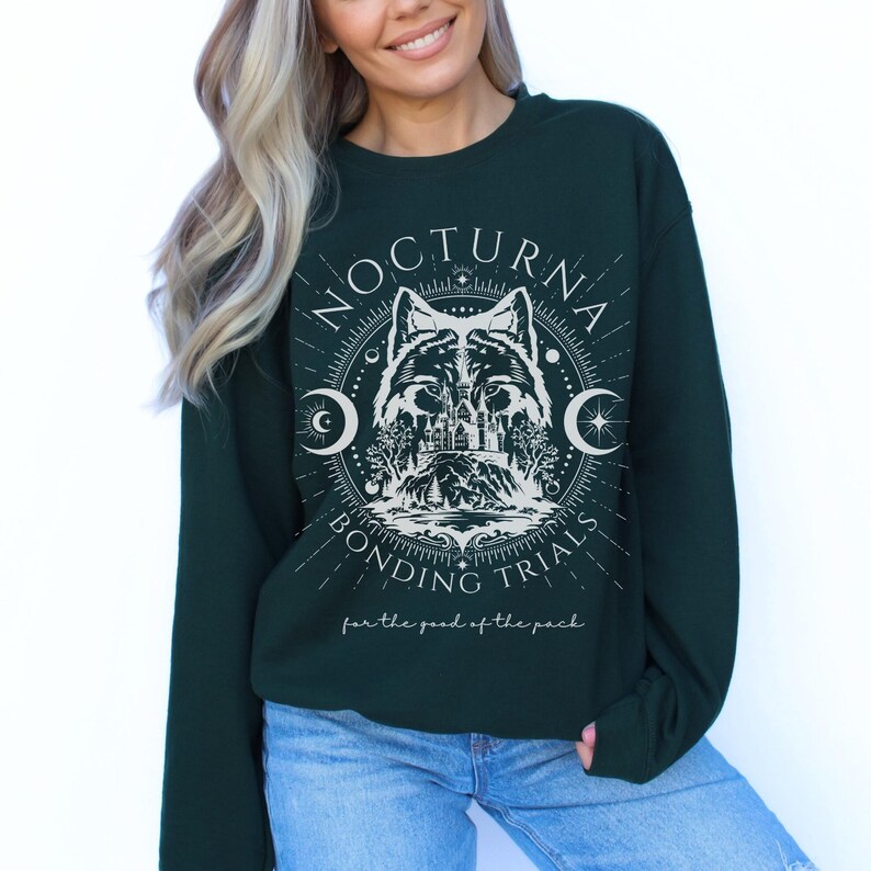 May include: Dark green sweatshirt with a white graphic featuring a wolf, castle, and crescent moons. The text reads "NOCTURNA BONDING TRIALS" and "for the good of the pack". The sweatshirt is a crew neck style.