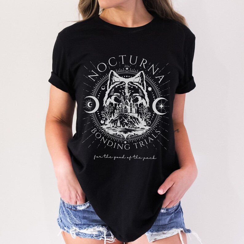 May include: Black t-shirt with a white graphic featuring a wolf, crescent moons, and the text "NOCTURNA BONDING TRIALS" in a circular design. Below the design is the text "for the good of the pack."