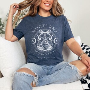 May include: Navy blue t-shirt with a white graphic featuring a wolf, crescent moons, and the text "NOCTURNA BONDING TRIALS". The shirt also includes the phrase "for the good of the pack".