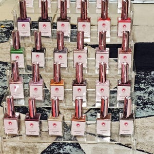 Puede incluir: Una exhibición de botellas de esmalte de uñas en una variedad de colores, desde rosas claros hasta rojos y marrones oscuros. Las botellas están dispuestas en un soporte transparente de varios niveles, mostrando los diferentes tonos disponibles. Cada botella tiene una tapa metálica de color oro rosa.