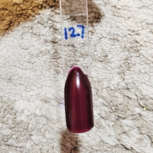 Puede incluir: Una muestra de esmalte de uñas en un color burdeos intenso. La muestra está en un palito de plástico transparente con el número "127" escrito en tinta azul. El fondo es una superficie texturizada de color claro.