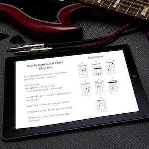 Könnte beinhalten: Ein schwarzes Tablet mit Gitarrenakkorddiagrammen und Anweisungen. Die Diagramme zeigen Dur-Akkorde. Eine rot-schwarze Gitarre befindet sich im Hintergrund, zusammen mit Plektren und einem Kabel.