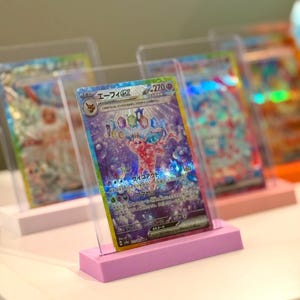 May include: Holographic trading cards in clear protective cases, displayed on pastel-coloured stands. The featured card has Japanese text and a vibrant illustration with purple and red hues. The stands are in shades of pink and peach.