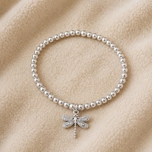 May include: A silver-toned beaded stretch bracelet featuring a small, detailed dragonfly charm. The bracelet is composed of numerous small, round, metallic beads strung together in a circular design with the dragonfly pendant hanging from the centre.