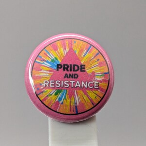 May include: A round, pink button with the words "PRIDE AND RESISTANCE" in black text. The button has a colorful, tie-dye-like design in the center, with a pink triangle. The button is on a white surface.