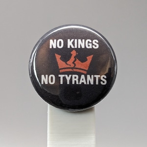 May include: Round black button with the text "NO KINGS NO TYRANTS" in white. A broken orange crown graphic is centered between the text. The button is on a white stand.