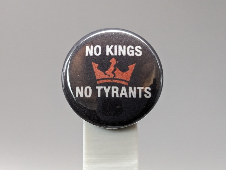 May include: Round black button with the text "NO KINGS NO TYRANTS" in white. A broken orange crown graphic is centered between the text. The button is on a white stand.