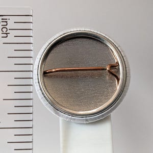 May include: A round button pin with a silver metal back and a copper-colored pin. The button is approximately 1 inch in diameter. The edge of the button is white with a subtle pattern.