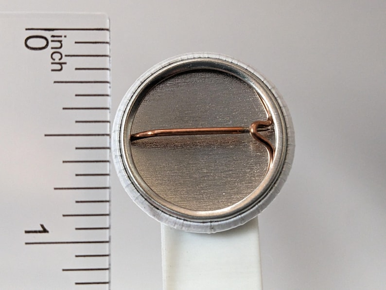 May include: A round button pin with a silver metal back and a copper-colored pin. The button is approximately 1 inch in diameter. The edge of the button is white with a subtle pattern.