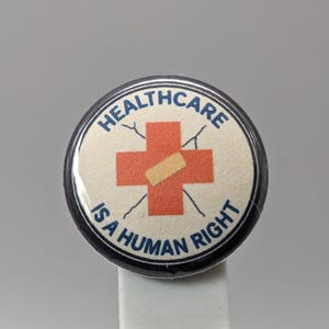 May include: Round button with a black border and the text "HEALTHCARE IS A HUMAN RIGHT" in blue. The center features a red cross with a bandage, symbolizing healthcare.