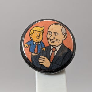 Puede incluir: Un botón redondo con una ilustración de dibujos animados de dos figuras políticas. Una figura sostiene a la otra. El botón tiene un borde negro y un fondo blanco.