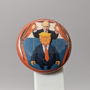 May include: A round button with a political cartoon illustration. The image depicts two figures: one with a blonde hairstyle and orange skin, wearing a suit and tie, and another figure behind him, giving a thumbs up.