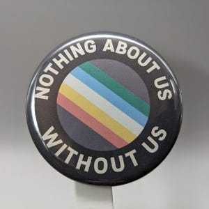May include: Round black button with the text "NOTHING ABOUT US WITHOUT US" in white. The center features a rainbow-colored stripe design. A simple, yet bold statement piece.