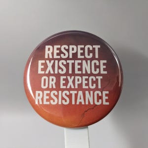 May include: A round button with the text "RESPECT EXISTENCE OR EXPECT RESISTANCE" in white letters. The button has a gradient background of orange, red, and brown. The button is attached to a white plastic stick.
