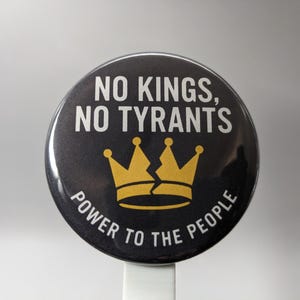 May include: A round, black button with white text reading "NO KINGS, NO TYRANTS" and "POWER TO THE PEOPLE." A gold crown with a crack down the middle is centered below the text. The button is on a white stick.