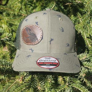 May include: An olive green baseball cap with a mesh back and a curved brim. The cap features a leather patch with a fly fishing design and a pattern of small, dark gray insects. The label reads "Imperial Mid-Crown Pro Fit Since 1936."