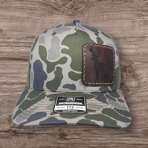 May include: A camouflage trucker hat with a brown leather patch featuring a silhouette of a deer. The hat has an adjustable strap and the text "Richardson 112 Original Trucker Adjustable" on the back.