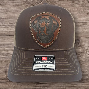 May include: Brown and tan trucker hat with a brown leather patch featuring a dog's head. The patch has stitching around the edges. The hat has a brown brim and a mesh back. The hat has a label that reads "EST. 1970 RICHARDSON. ORIGINAL 112 TRUCKER ADJUSTABLE".