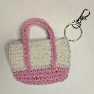 May include: A handmade crochet keychain in the shape of a small tote bag. The bag is white with a pink base and pink handles. A silver-colored metal clasp and ring are attached to the bag.
