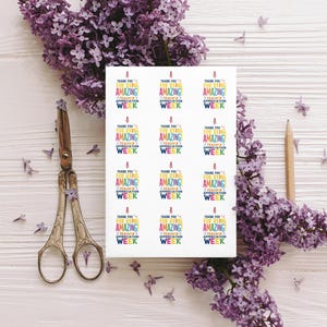 Peut inclure: Une feuille d'autocollants avec le texte "Thank You For Being Amazing! Teacher Appreciation Week" en plusieurs couleurs. Les autocollants sont entour&eacute;s de fleurs de lilas violettes, d'une paire de ciseaux et d'un crayon sur une surface en bois blanc.