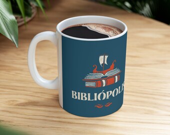 Può includere: Una tazza in ceramica bianca con sfondo blu e un'illustrazione in stile vintage di una pila di libri con una vela di barca in cima. Il testo "BIBLIÓPOLIS" è stampato in bianco sotto l'illustrazione.