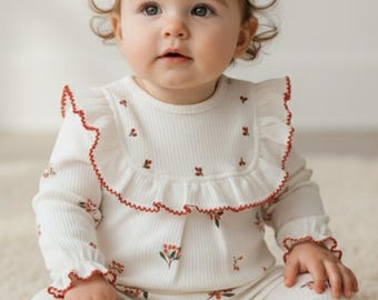 May include: A baby wearing a white ribbed knit outfit with a floral pattern and red trim. The outfit includes a long-sleeved top with a ruffled collar and cuffs, and matching footed pants. A white headband with a bow completes the ensemble.