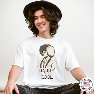 May include: White t-shirt with a graphic of a man with an afro, sunglasses, and suspenders. The text "DADDY COOL" is printed below the graphic. The shirt is worn by a person wearing a black hat.