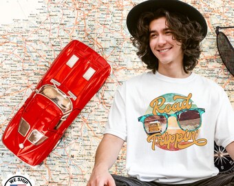 Op de afbeelding: Een rode speelgoedauto en een wit t-shirt met de tekst "Road Trippin" erop. Het t-shirtontwerp toont een zonnebril met een schilderachtig uitzicht en een gele bus. De achtergrond is een kaart.