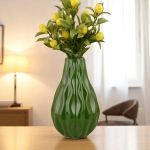 May include: A green ceramic vase with a textured, wavy design holds a bouquet of artificial lemons and green leaves. The vase is on a wooden table, with a lamp and artwork in the background.