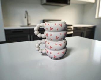 May include: Two stacked white ceramic mugs with a heart pattern. Each mug has a unique handle and is decorated with small red hearts. The mugs are on a white surface, with a kitchen in the background.