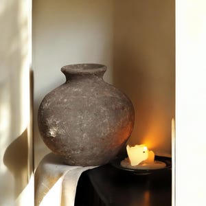 May include: A large, round, textured, brown vase with a narrow neck sits on a white cloth. A lit candle in a black dish is next to the vase. The scene is lit with warm, natural light, creating shadows.