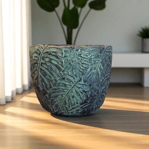 Può includere: Un grande vaso rotondo color verde acqua con una superficie strutturata con un motivo ripetuto di foglie di monstera. Il vaso è realizzato in un materiale resistente ed è adatto per uso interno o esterno. Il vaso è vuoto e pronto per la semina.