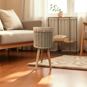 Op de afbeelding: Een grijze geweven plantenbak met een decoratieve rand staat op drie houten poten. De plantenbak staat in een woonkamer met een bank, stoel en vloerkleed. De kamer heeft hardhouten vloeren en natuurlijk licht.