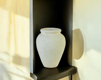 May include: A textured, off-white vase with a wide mouth and a rounded body. The vase sits on a black shelf against a light-colored wall. The vase has a rough, aged appearance, suggesting a rustic or antique style.