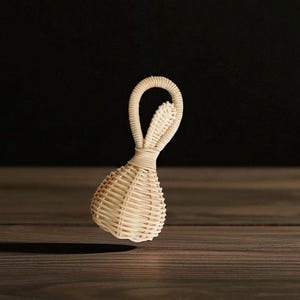 May include: A woven rattan baby rattle in the shape of a bunny with a rounded body and looped handle. The rattle is light beige and appears to be floating above a dark wooden surface. The background is a dark, solid color.