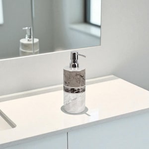 Puede incluir: Un dispensador de jabón con diseño de mármol y bomba plateada. El dispensador cilíndrico está sobre una encimera blanca. El entorno del baño incluye un espejo y un mueble blanco.