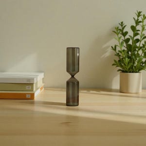 Pode incluir: Um vaso de vidro cinza em forma de ampulheta está sobre uma superfície de madeira. O vaso tem uma cintura estreita e uma parte superior e inferior alargadas. À direita, há uma pequena planta em vaso com folhas verdes. Três livros estão empilhados à esquerda.