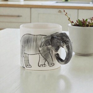 May include: White ceramic mug with a hand-painted elephant design. The elephant's body wraps around the mug, with the trunk forming the handle. The mug is on a white table, next to a small white pot with a plant.