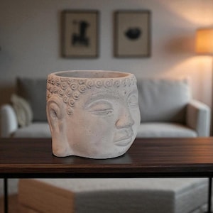 Puede incluir: Una maceta con forma de cabeza de Buda gris claro hecha de hormigón. La maceta tiene los ojos cerrados y rasgos faciales detallados. La maceta decorativa es adecuada para uso en interiores o exteriores y está colocada sobre una mesa marrón oscuro.