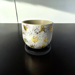Può includere: Un vaso di ceramica con un esterno bianco e nero maculato, adornato con accenti di foglia d'oro. Il vaso poggia su un piccolo sottobicchiere rotondo nero. L'interno del vaso è di colore beige chiaro.