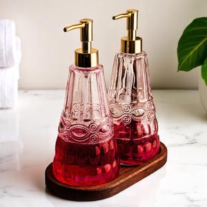 Pode incluir: Dois dispensadores de sabonete de vidro rosa com bombas douradas estão em uma bandeja de madeira. Os dispensadores têm um design ornamentado e texturizado e são preenchidos com um líquido vermelho. O fundo inclui toalhas brancas e uma planta.