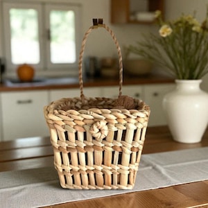 Könnte beinhalten: Ein quadratischer Weidenkorb mit Henkel, hergestellt aus Naturfasern. Der Korb hat einen dekorativen Knoten auf der Vorderseite und steht auf einem Holztisch mit einem neutralen Tischläufer. Eine weiße Vase mit Blumen ist im Hintergrund.