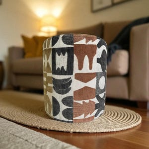 Può includere: Un pouf cilindrico con un motivo geometrico nei toni del nero, marrone e crema. Il pouf è posizionato su un tappeto rotondo in juta intrecciata. Lo sfondo comprende un divano e un pavimento in legno.