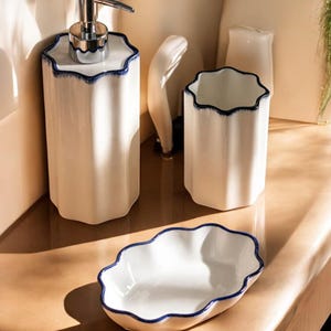 May include: A white ceramic bathroom set with a wavy blue trim. The set includes a soap dispenser, a tumbler, and a small dish. The items are arranged on a light-colored surface, with shadows adding depth to the scene.