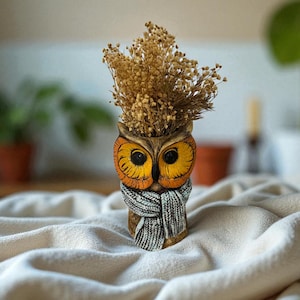 May include: A hand-painted owl-shaped planter with a scarf, holding dried flowers. The owl has large yellow eyes and orange and brown coloring. The planter is sitting on a white, textured surface.