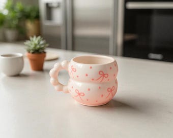 May include: A light pink ceramic mug with a unique double-layered design. The mug features a handle made of connected circles and is decorated with small pink bows and dots. The mug is sitting on a white surface.