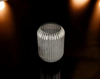 Puede incluir: Un jarrón cilíndrico de vidrio transparente con detalles acanalados verticales. El jarrón tiene una parte superior redondeada y una superficie reflectante, adecuado para la decoración del hogar. El fondo es negro con focos brillantes.