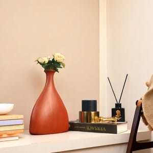 May include: A terracotta-colored vase holding white roses sits on a white shelf next to a stack of books and a white bowl. A black diffuser and a book with the word "VERSACE" are also on the shelf.