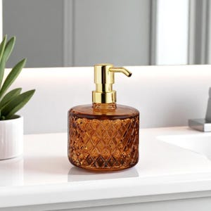 Può includere: Dispenser per sapone in vetro ambrato con motivo a diamante e pompa color oro. Il dispenser cilindrico è posto su una superficie bianca. Una piccola pianta in vaso a sinistra.